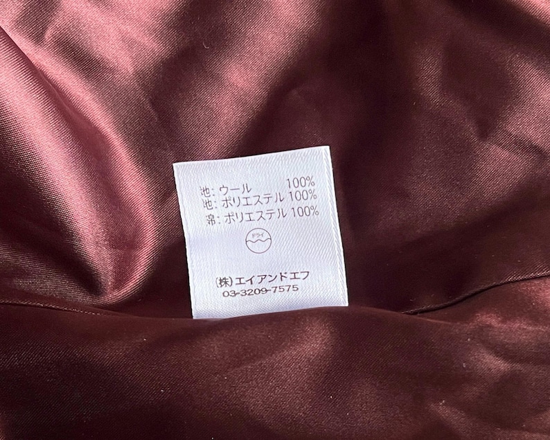 Op de afbeelding: Close-up van een bordeauxrode stoffen binnenkant met een wit label. Het label heeft Japanse tekst die de samenstelling van de stof aangeeft: 100% wol, 100% polyester, en een bedrijfsnaam en telefoonnummer.