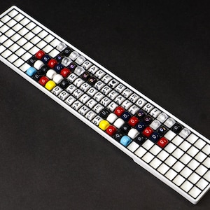 May include: A rectangular white grid board featuring rows of colorful plastic beads. Some beads are printed with letters forming the words RAVE, SPROUT, BREATHE, MINDFUL, SMILE, and DREAMER. The beads are arranged in a pattern across the grid surface.