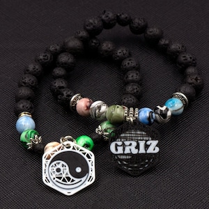 May include: Two black lava rock bracelets with colorful beads and charms. One charm is a yin yang symbol and the other says "GRIZ".
