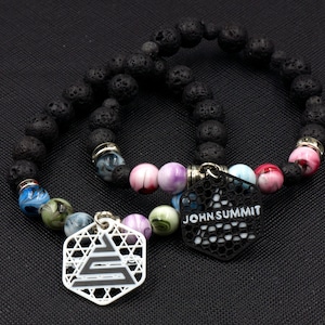 May include: Two black lava rock bracelets with colorful glass beads and metal charms. One charm is a hexagon with the text "JOHN SUMMIT" and the other is a white hexagon with a geometric design.