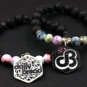 May include: Two black lava rock bracelets with colorful beads and charms. One charm says "daily bread" and the other has a black and white design with the letters "DB".