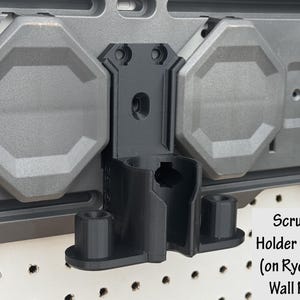 May include: Black scrubber holder designed for Ryobi Link wall rails. The holder is mounted on a gray wall panel with octagonal gray accents. The text "Scrubber Holder in Black (on Ryobi Link Wall Rails)" is visible.