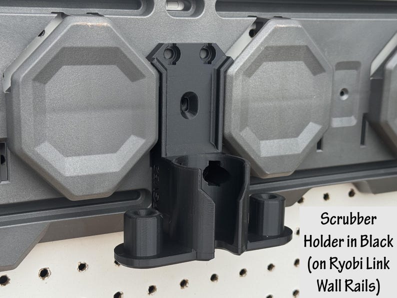 May include: Black scrubber holder designed for Ryobi Link wall rails. The holder is mounted on a gray wall panel with octagonal gray accents. The text "Scrubber Holder in Black (on Ryobi Link Wall Rails)" is visible.