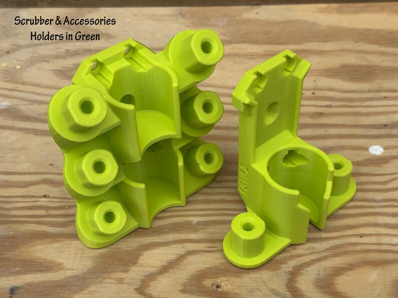 May include: Two bright green scrubber holders with multiple circular openings and a curved design. The text "Scrubber & Accessories Holders in Green" is at the top left. The holders are on a wooden surface.