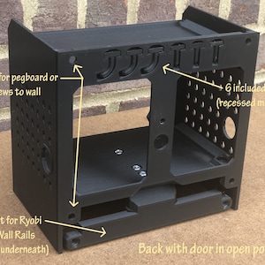 May include: Black plastic wall mount with a door that opens. It has six pegs and four holes for mounting to a wall. The mount is designed to hold Ryobi Link Wall Rails.