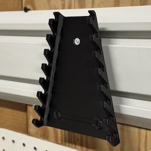 Puede incluir: Un organizador de llaves negro montado sobre una superficie blanca y de madera. El organizador es triangular, con múltiples ranuras para sujetar llaves de varios tamaños. Un solo perno asegura el organizador a la pared.