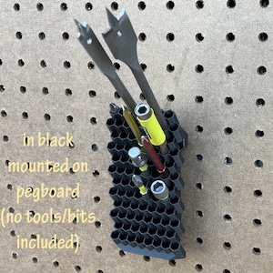 Könnte beinhalten: Ein schwarzer, wabenförmiger Werkzeug-Organizer, der an einer Lochwand montiert ist. Der Organizer ist mit verschiedenen Werkzeugen gefüllt, darunter Bohrer und Schraubendreher. Der Text "in black mounted on pegboard (no tools/bits included)" ist im Bild sichtbar.
