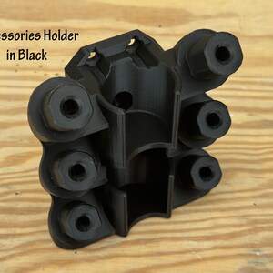 May include: Black accessories holder with multiple circular openings and a central cylindrical cavity. The text "Accessories Holder in Black" is visible. The holder is made of a matte black material and sits on a light brown wooden surface.