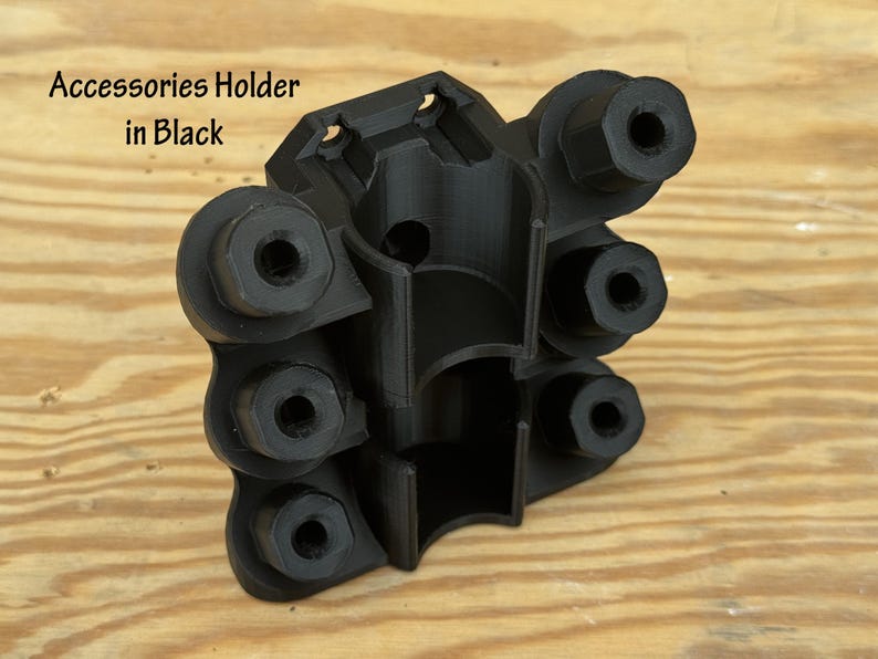 May include: Black accessories holder with multiple circular openings and a central cylindrical cavity. The text "Accessories Holder in Black" is visible. The holder is made of a matte black material and sits on a light brown wooden surface.
