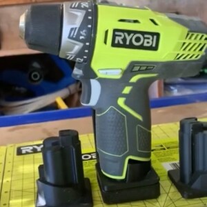 May include: A green and black Ryobi drill with a black charging stand. The drill has a black handle and a yellow body with the word "RYOBI" printed on it.