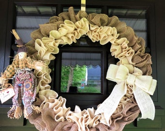 May include: A burlap wreath with a large white bow and a small scarecrow figurine with the text "I Love Fall" hanging on a brown door.