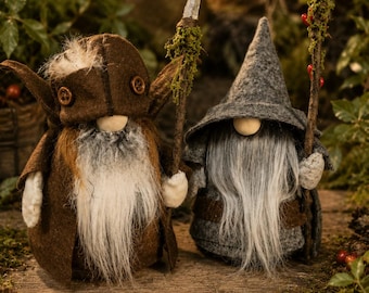 May include: Two plush gnome figures with long white beards. The left gnome wears a brown felt hat with buttons and holds a staff topped with a clear crystal. The right gnome wears a grey felt hat and holds a staff decorated with moss and red berries.
