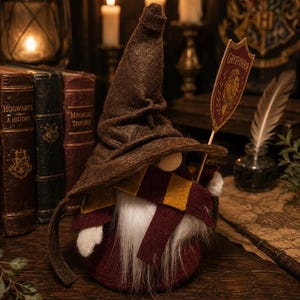 Könnte beinhalten: Eine Zwergenfigur mit einem hohen braunen Filzhut und einem kastanienbraun-gold gestreiften Schal. Der Zwerg hat einen langen weißen Bart und hält ein kleines Schild an einem Stab mit der Aufschrift Gryffindor und einem Löwenemblem in Rot und Gold.