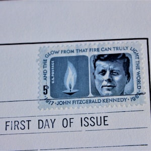 Pode incluir: Um selo postal comemorativo azul e branco com um retrato de John F. Kennedy e uma vela acesa com o texto "And the glow from that fire can truly light the world." e "1917 John Fitzgerald Kennedy 1963"