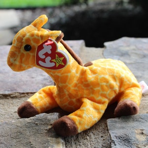 Pode incluir: Um brinquedo de pelúcia de girafa amarelo e marrom com uma etiqueta vermelha Beanie Baby que diz "Beanie Babies" e tem uma estrela.