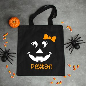 May include: Black canvas tote bag with a white jack-o'-lantern face with a bow, eyelashes, and the name Peyton printed in orange.