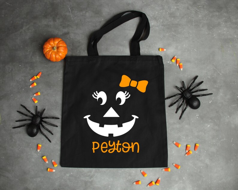 May include: Black canvas tote bag with a white jack-o'-lantern face with a bow, eyelashes, and the name Peyton printed in orange.