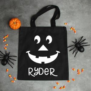May include: Black canvas tote bag with a white jack-o'-lantern face and the name "Ryder" printed on the front. The bag is surrounded by candy corn and toy spiders.