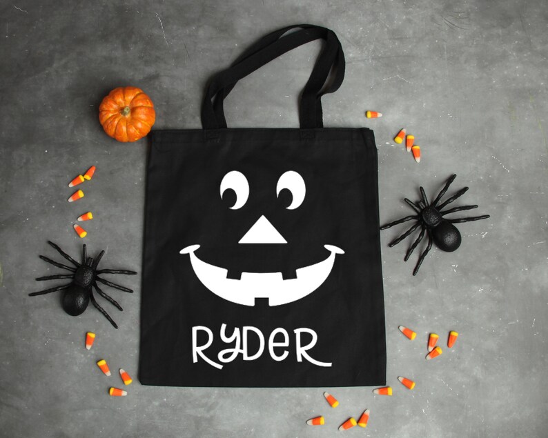 May include: Black canvas tote bag with a white jack-o'-lantern face and the name "Ryder" printed on the front. The bag is surrounded by candy corn and toy spiders.