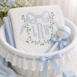 Peut inclure: Un panier en osier blanc contient une couverture pour bébé, un hochet et un bloc de jouet. La couverture blanche est brodée d'un nœud bleu et d'un monogramme. Le hochet est blanc à pois bleus et un ruban bleu. Un bloc de jouet bleu est également présent.