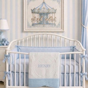 Op de afbeelding: Een witte wieg met blauwe beddengoed en een bijpassende quilt met de naam "HENRY" geborduurd. Een ingelijste foto van een carrousel hangt boven de wieg. De kamer heeft blauw-wit gestreept behang en gordijnen.