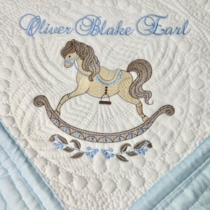 Puede incluir: Manta de bebé blanca con borde azul y diseño de caballo balancín bordado. La manta presenta el nombre "Oliver Blake Earl" en escritura azul. El caballo balancín es beige y marrón, con detalles azules.