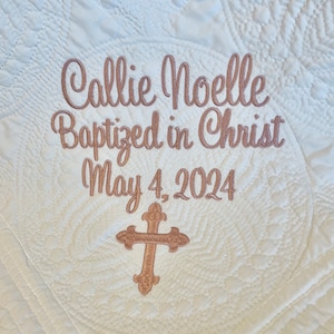 May include: White quilted fabric with embroidered text "Callie Noelle Baptized in Christ May 4, 2024" and a gold cross.