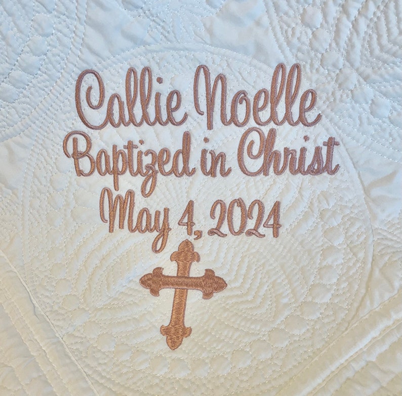 May include: White quilted fabric with embroidered text "Callie Noelle Baptized in Christ May 4, 2024" and a gold cross.