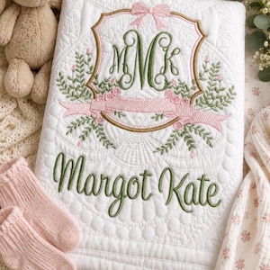 May include: A white quilted baby blanket featuring an embroidered crest with a pink bow, green floral accents, and the monogram MMK. The name Margot Kate is embroidered in green script below the crest. The blanket has a textured, scalloped pattern.