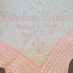 May include: A white and pink quilted baby blanket with the text "Caroline Lucille Baptized in Christ April 21, 2024" embroidered in pink. A pink cross is embroidered in the centre of the blanket.
