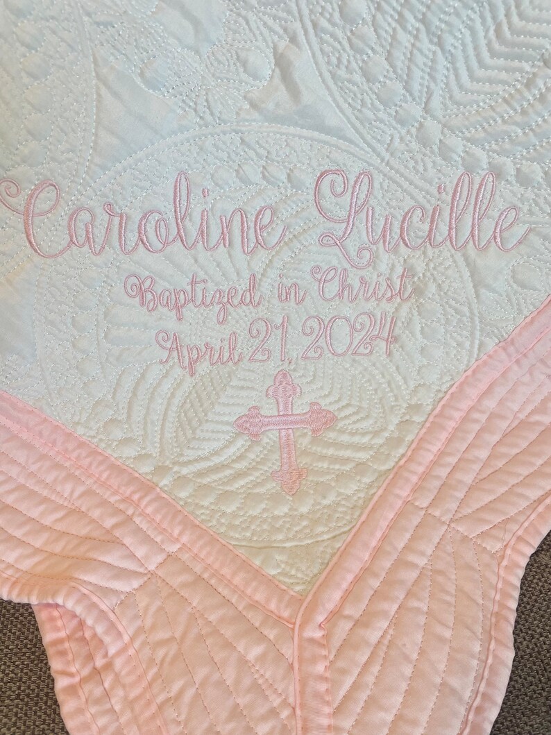 May include: A white and pink quilted baby blanket with the text "Caroline Lucille Baptized in Christ April 21, 2024" embroidered in pink. A pink cross is embroidered in the centre of the blanket.