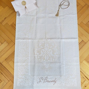 Puede incluir: Alfombra de oraci&oacute;n blanca con un dise&ntilde;o bordado en oro y flecos. La alfombra tiene una peque&ntilde;a bolsa blanca con un borla dorada y una cadena de oraci&oacute;n dorada.