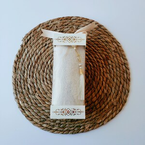 May include: A white box with gold trim and a white fabric inside. The box is sitting on a brown woven mat.
