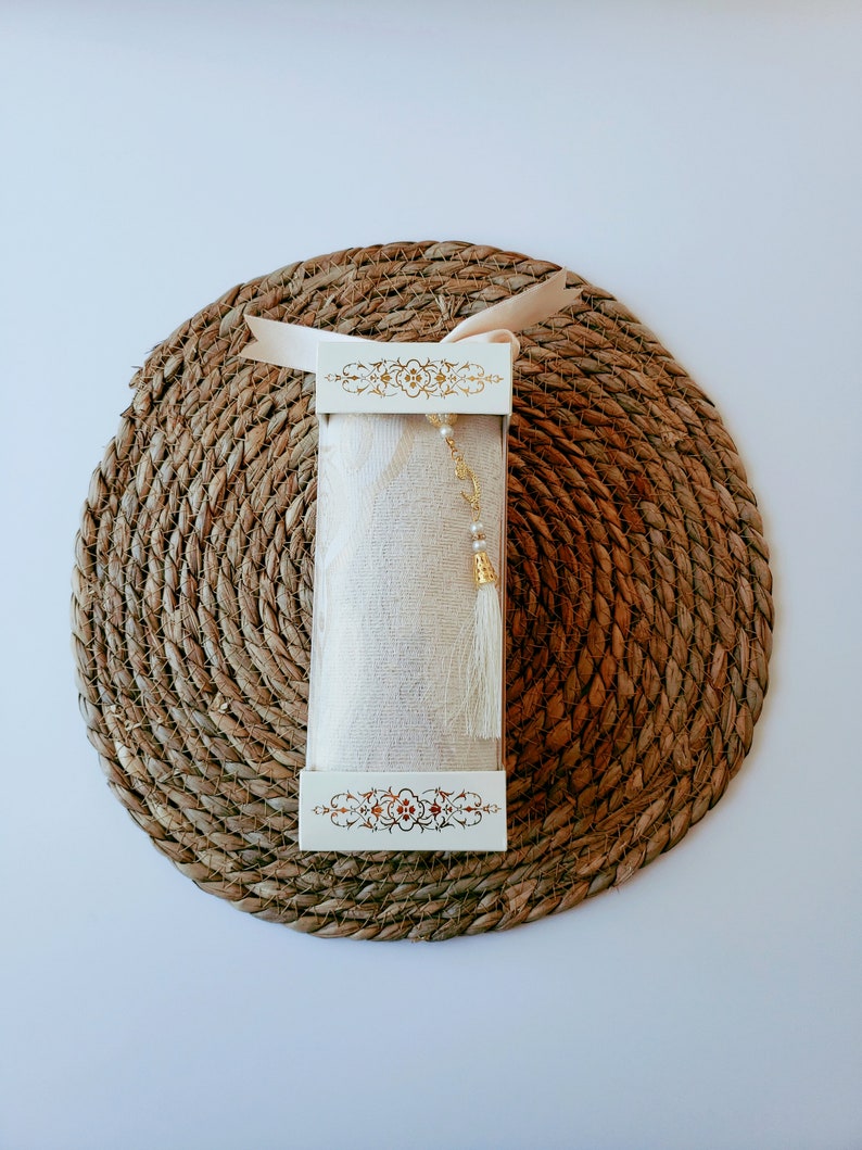 May include: A white box with gold trim and a white fabric inside. The box is sitting on a brown woven mat.