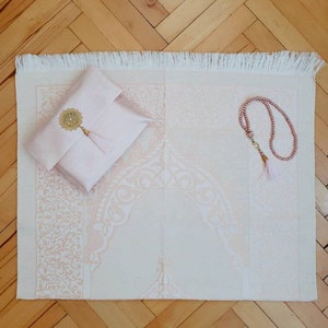 Puede incluir: Una alfombra de oraci&oacute;n rosa con flecos blancos, una alfombra de oraci&oacute;n rosa con un borla dorada y un collar de cuentas de oraci&oacute;n rosa.