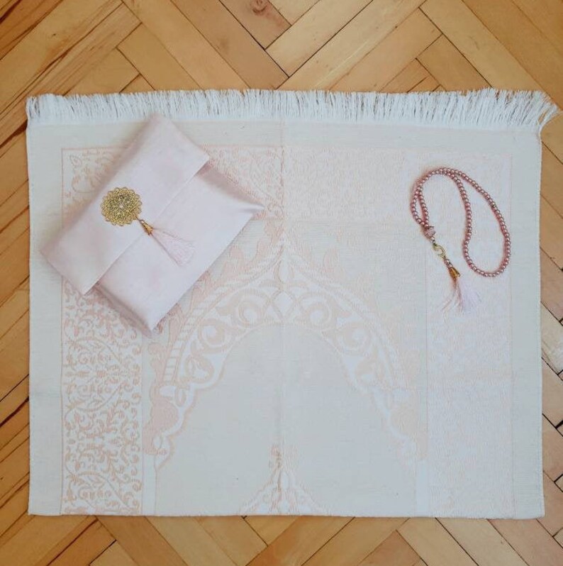 Puede incluir: Una alfombra de oraci&oacute;n rosa con flecos blancos, una alfombra de oraci&oacute;n rosa con un borla dorada y un collar de cuentas de oraci&oacute;n rosa.