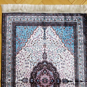 Op de afbeelding: Een gebedskleed met een witte achtergrond en ingewikkelde bloemmotieven in tinten blauw, roze en bruin. Het kleed heeft een zwarte rand met een decoratief ontwerp en een franje aan de bovenrand.
