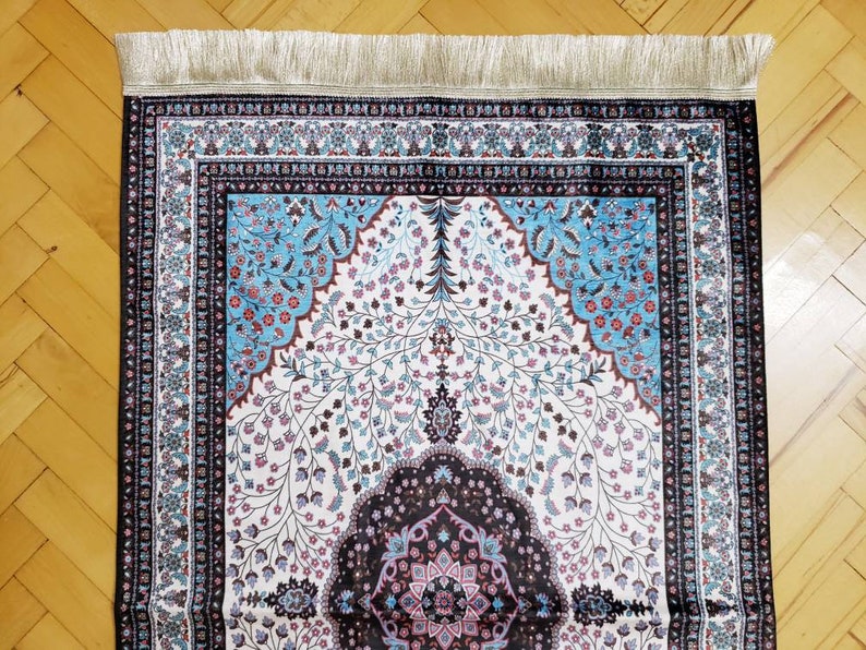 Op de afbeelding: Een gebedskleed met een witte achtergrond en ingewikkelde bloemmotieven in tinten blauw, roze en bruin. Het kleed heeft een zwarte rand met een decoratief ontwerp en een franje aan de bovenrand.