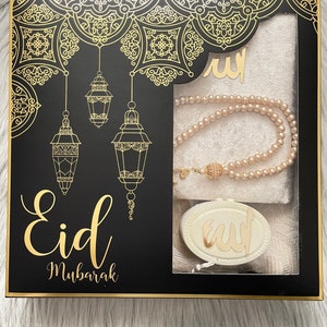 May include: A black gift box with gold detailing, featuring the words "Eid Mubarak". The box contains a prayer mat, a pearl necklace, and a decorative pendant. The box has a gold border and decorative patterns.