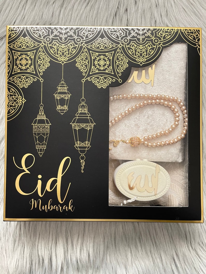 May include: A black gift box with gold detailing, featuring the words "Eid Mubarak". The box contains a prayer mat, a pearl necklace, and a decorative pendant. The box has a gold border and decorative patterns.