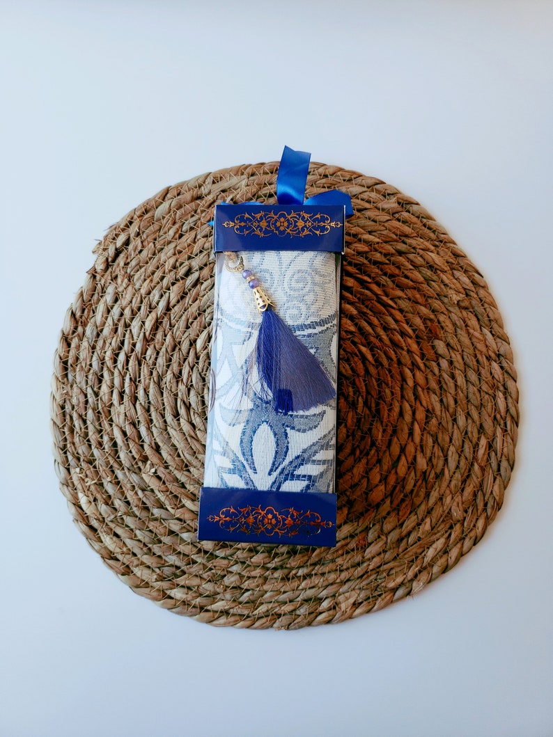 May include: A blue and white prayer rug with a tassel, packaged in a blue box with gold accents. The rug is on a woven straw mat.