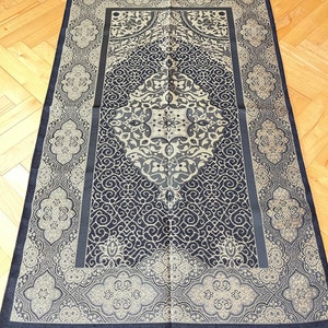 May include: A navy blue and beige prayer rug with a detailed, symmetrical pattern. The rug has a central diamond design and decorative borders. The edges are finished with a navy fringe, and the rug is rectangular in shape.