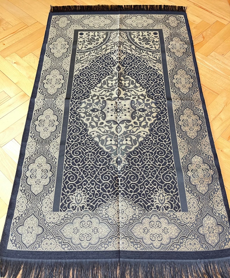 May include: A navy blue and beige prayer rug with a detailed, symmetrical pattern. The rug has a central diamond design and decorative borders. The edges are finished with a navy fringe, and the rug is rectangular in shape.