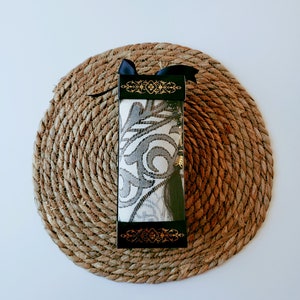 May include: A black and gold gift box with a white and gray patterned fabric inside. The box is tied with a black ribbon and is sitting on a woven straw mat.