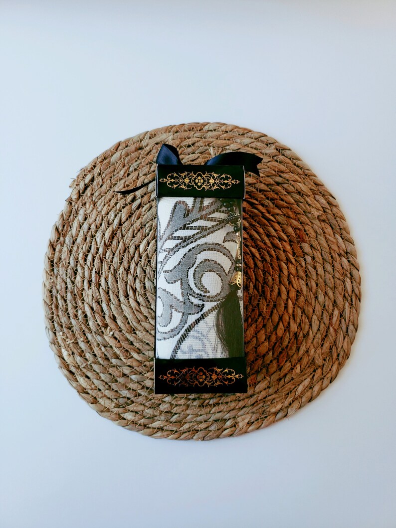 May include: A black and gold gift box with a white and gray patterned fabric inside. The box is tied with a black ribbon and is sitting on a woven straw mat.
