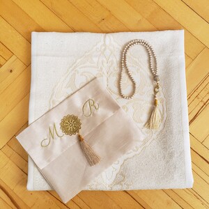 Puede incluir: Una alfombra de oraci&oacute;n blanca con un dise&ntilde;o bordado en oro. Una alfombra de oraci&oacute;n beige m&aacute;s peque&ntilde;a con bordado dorado y las iniciales "M & R" est&aacute; encima de la alfombra m&aacute;s grande. Un collar de cuentas de oraci&oacute;n blanco con un borla dorado est&aacute; en la alfombra m&aacute;s grande.