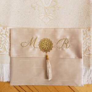 Puede incluir: Una alfombra de oraci&oacute;n beige con un borla dorada y un medall&oacute;n decorativo dorado con las iniciales "M" y "R" bordadas en la alfombra.