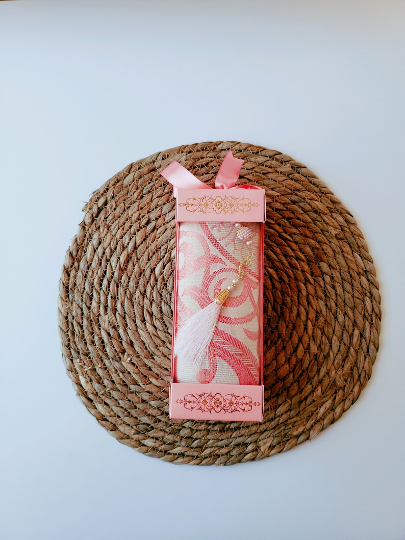 May include: A pink prayer rug with a white tassel and gold beads is packaged in a pink box with a floral design. The box is sitting on a woven straw mat.