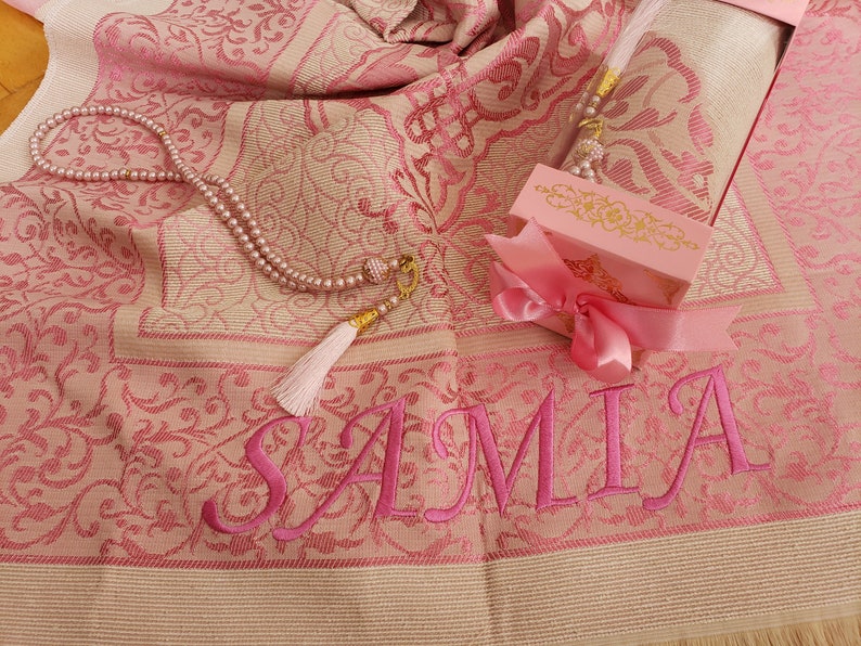 May include: A pink prayer rug with the name "SAMIA" embroidered on it. The rug is folded and a pink gift box with a pink ribbon is on top of it. A pink prayer bead necklace with a tassel is on the rug.