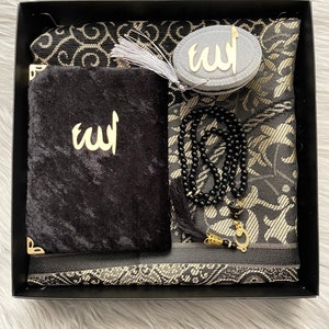May include: A black gift box containing a prayer mat, a velvet pouch, a prayer bead, and a compass. The prayer mat has a black and gold floral pattern. The pouch and compass feature the word "Allah" in gold.